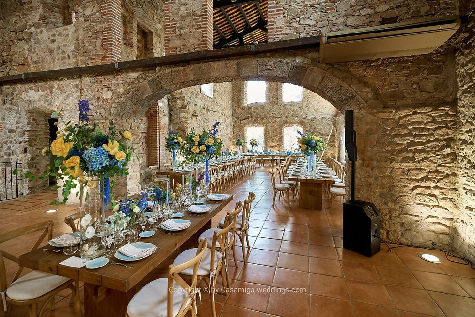 Indoor reception space prepared as a practical weather backup at a Spanish wedding venue. Elegant wedding reception in a stone hall with long wooden tables, blue and yellow floral centerpieces, and set tables under arches.