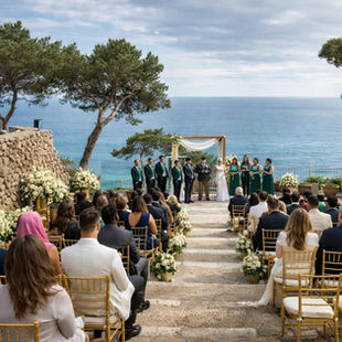 Costa Brava wedding 2026 sea view ceremony Casamiga Weddings. Outdoor wedding ceremony by the ocean with guests seated on stone steps. The bride and groom stand under an arch. Bridesmaids in green dresses.
