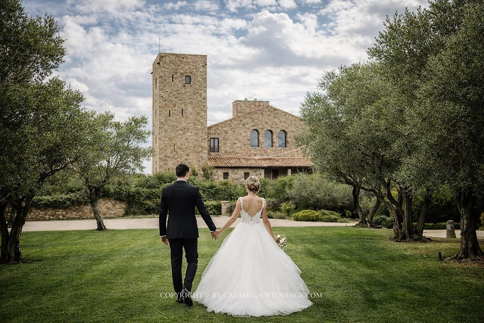 Bride and groom at a coastal ceremony venue in Spain planned by Casamiga Weddings. Bride and groom walk hand in hand across a green lawn toward a stone villa under cloudy skies; copyright watermark below.