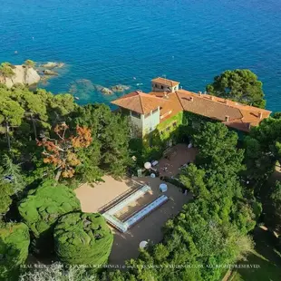 Aerial view of a Mediterranean villa surrounded by lush gardens in Casa de Santa Clotilde, Catalonia, Spain, featured in the Casamiga Weddings portfolio. Aerial view of a terracotta-roofed seaside villa amid lush trees, overlooking the bright blue sea; wedding watermark text at the bottom.