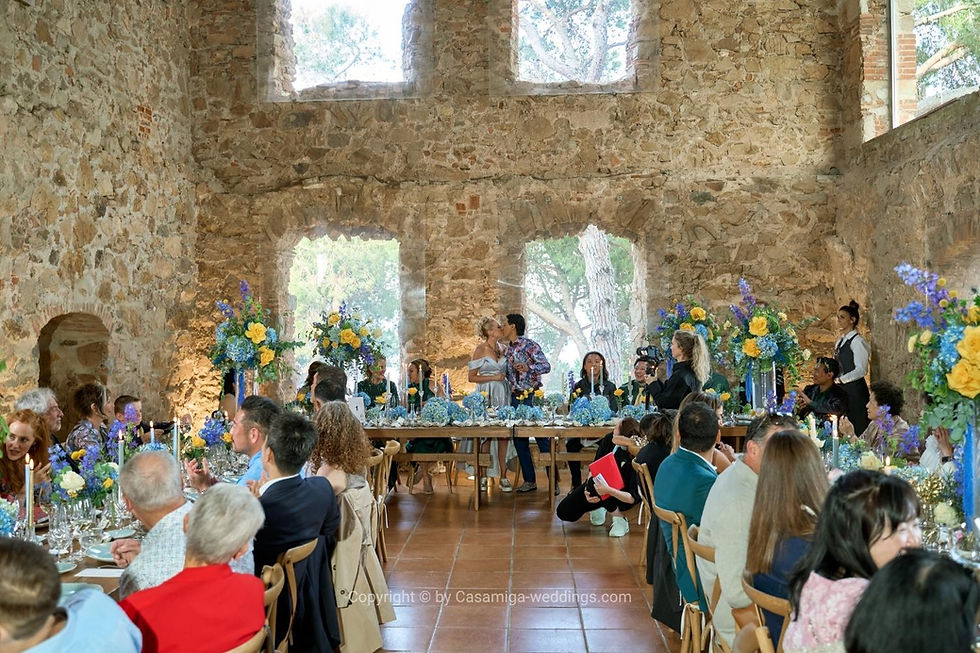 Private estate wedding in Girona with full planning by Casamiga Weddings. The wedding couple kisses in a rustic stone hall, surrounded by guests and blue-yellow floral centerpieces.