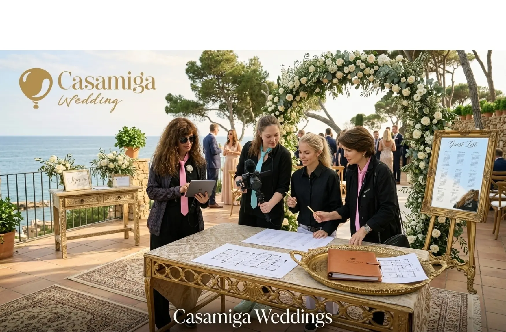 Casamiga Weddings planner reviewing the full-day timeline for a Casa de Santa Clotilde wedding with an international couple. Foto: Personas/vestuario mejorados mediante IA. Wedding planners confer at a seaside Casamiga Weddings venue, with floral arch, guest list, and tables by the ocean.