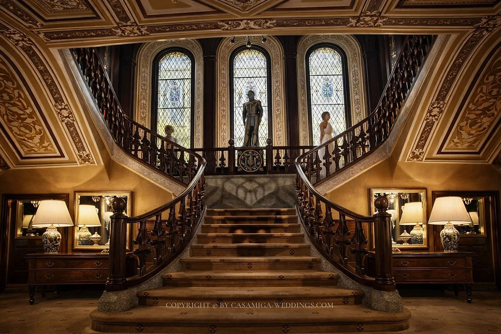 Historic architecture and a Mediterranean garden setting at Bell Recó near Barcelona. Grand ornate staircase in a mansion with stained-glass windows, statues, warm lamps, and a CASAMIGA-WEDDINGS.COM watermark.