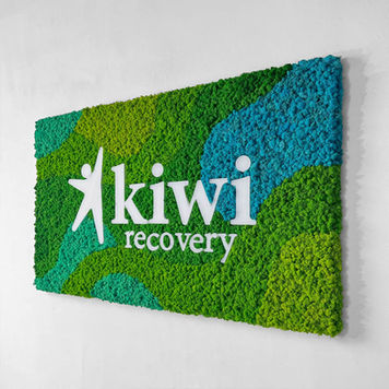 Patterned reindeer moss wall with a custom Kiwi Recovery logo in blue and green tones, creating a vibrant branded biophilic feature wall.
