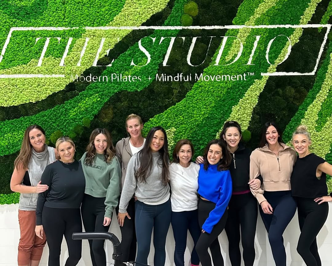 The Studio pilates team posing in front of seamless moss wall with "Modern Pilates + Mindful Movement" branding in fitness studio
