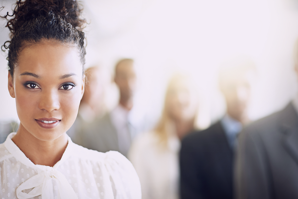 Smiling woman in white blouse in focus, standing confidently. Blurred group of people in business attire behind her. Bright, soft-lit setting.