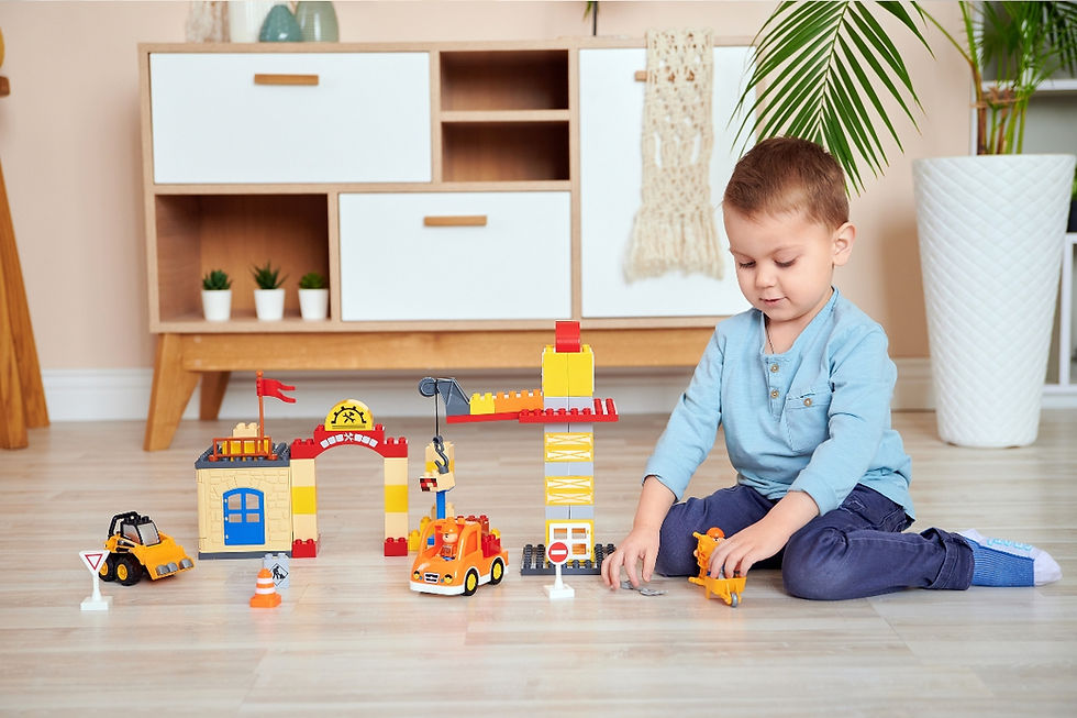 Ein kleines Kind spielt konzentriert mit LEGO-Steinen in einem Wohnzimmer, umgeben von Spielsachen und Möbeln.