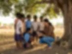 Outdoor photography. A woman educates a group of schoolchildren under a tree, demonstrating an object in a natural, rural setting.
