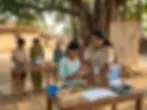Outdoor education photography of a teacher guiding children on a solar power project in a rural village. Students are actively engaged with electronic components and learning materials.