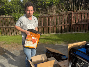 Harley standing outdoors beside open boxes of new equipment, holding a bag of welding supplies in a backyard setting.