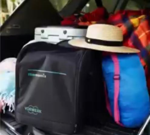 Car trunk packed with a straw hat, blue-pink sleeping bag, black Vorwerk bag, and colorful blanket. Suitcase visible, outdoor light. Travelling with Thermomix