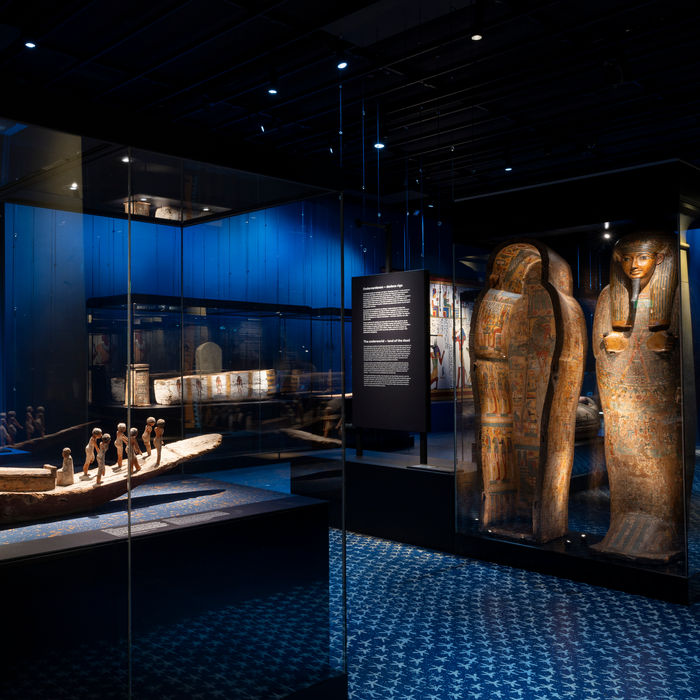 The traveling museum exhibit Ancient Egypt: in the foreground, a wooden model of a funerary boat with small figures in a glass display case. To the right, several decorated wooden anthropoid sarcophagi stand upright. The room is dimly lit with blue walls and floor, featuring large-scale reproductions of Egyptian wall paintings in the background.