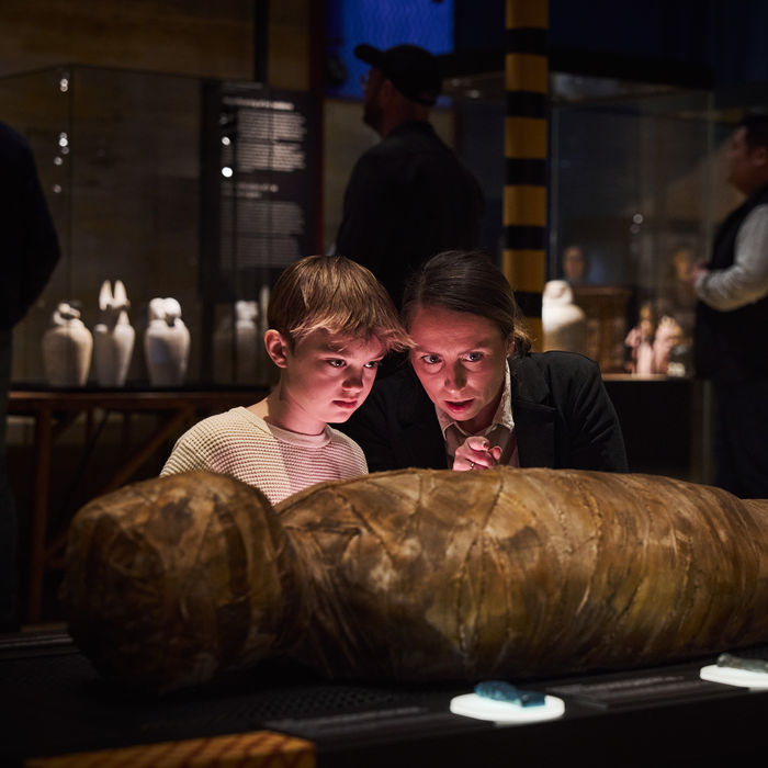 A woman and a young boy lean in closely to examine the interactive mummy wrapped in brown linen. They can virtually trigger the protective amulets hidden within the mummy's wrappings Other visitors and canopic jars are visible in the soft-focus background.