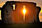 Close-up of Stonehenge at sunrise. The bright sun glows through a narrow gap between massive stone trilithons, casting a warm orange light over the ancient monument.