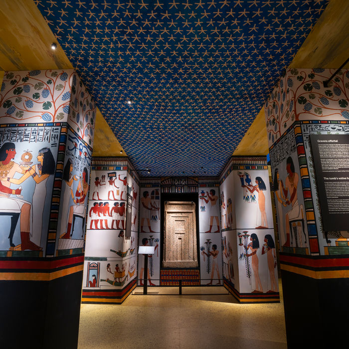 Full-scale walk-in replica of an ancient Egyptian offering hall. The walls are covered in colorful, detailed paintings of figures and hieroglyphs, and the ceiling is painted deep blue with a dense pattern of yellow stars. An original ancient Egyptian false door stands at the far end of the narrow corridor.