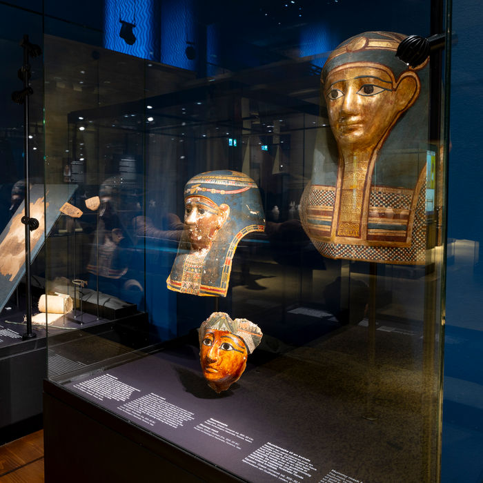 Several ancient Egyptian mummy masks from the Ptolemaic Period displayed in glass museum cases. One large gilded mask with a serene expression and painted details stands prominently in the foreground, alongside smaller colorful masks and fragments.