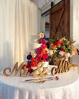 Four tiered wedding cake decorated with brightly colored flowers and a gold Mr & Mrs sign in front of it
