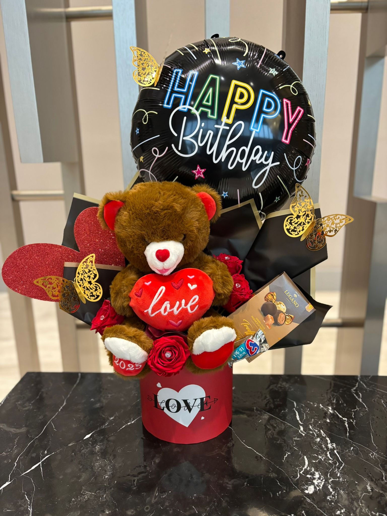 Teddy bear holding heart with 'LOVE', Happy Birthday balloon, floral bouquet.