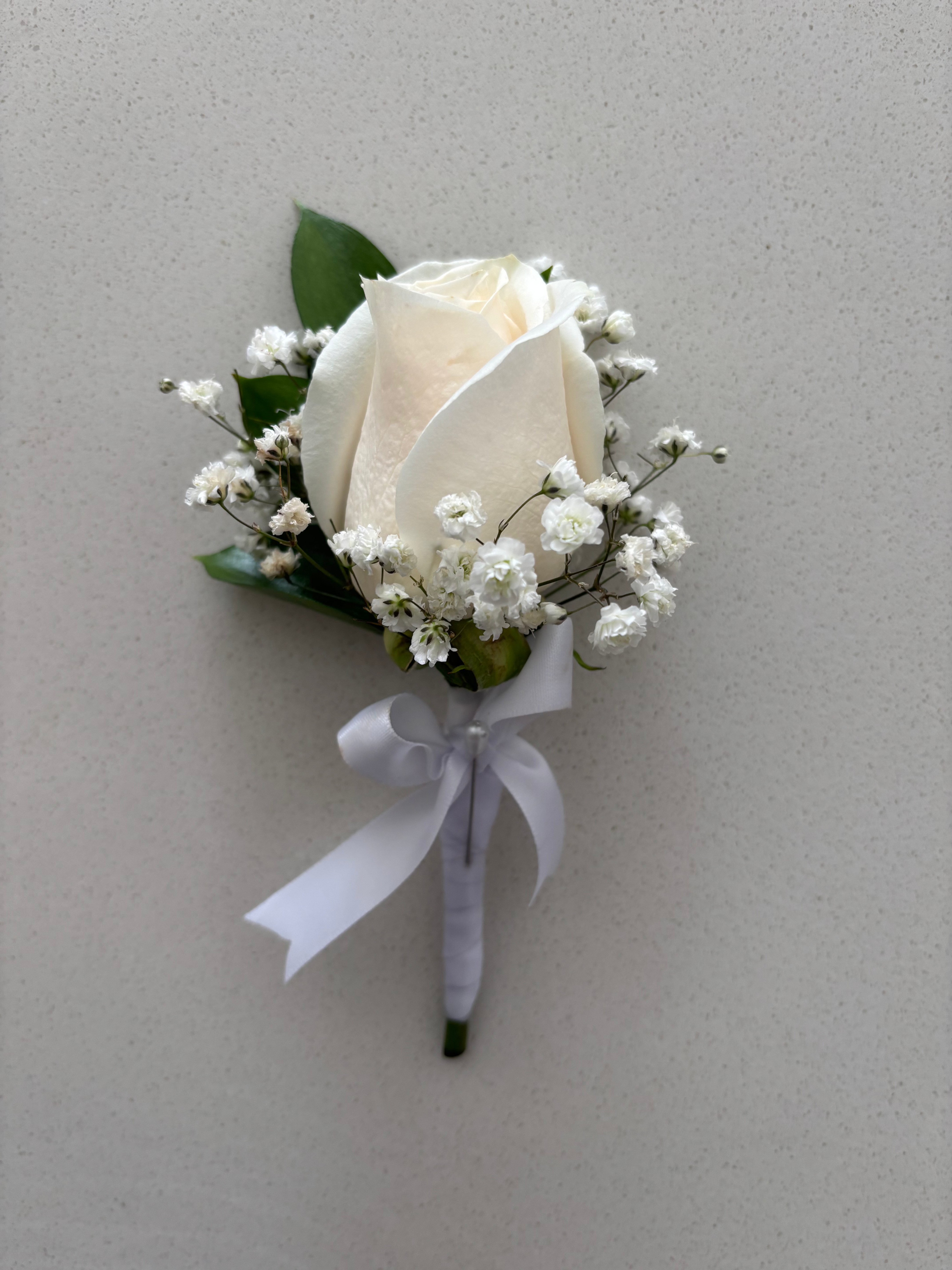 Single white rose boutonniere with baby's breath and ribbon on neutral background. Doral Blooms