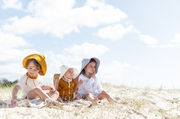 three children wearing hats sit on a sandy beach