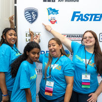 Lady Jacket Innovators in teal team shirts pointing the the Sporting KC logo on the step and repeat banner