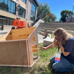team member in navy team shirt spray painting her wooden prototype outside in the grass