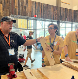two team members in gold team shirts standing at a table holding wood next to a purdue tool tech