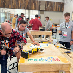 tool tech gregg holding a hammer and screw driver working on team teams wooden prototype as they stand next to him in their grey team shirts