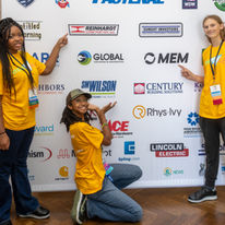 Three student team members from WomEngineers pointing to their sponsor logos on the step and repeat banner
