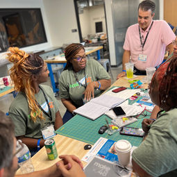 a mentor talking with a team sitting at their team table in green team shirts