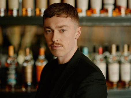 Man with a mustache in a black blazer stands before blurred liquor bottles in a bar, looking serious.