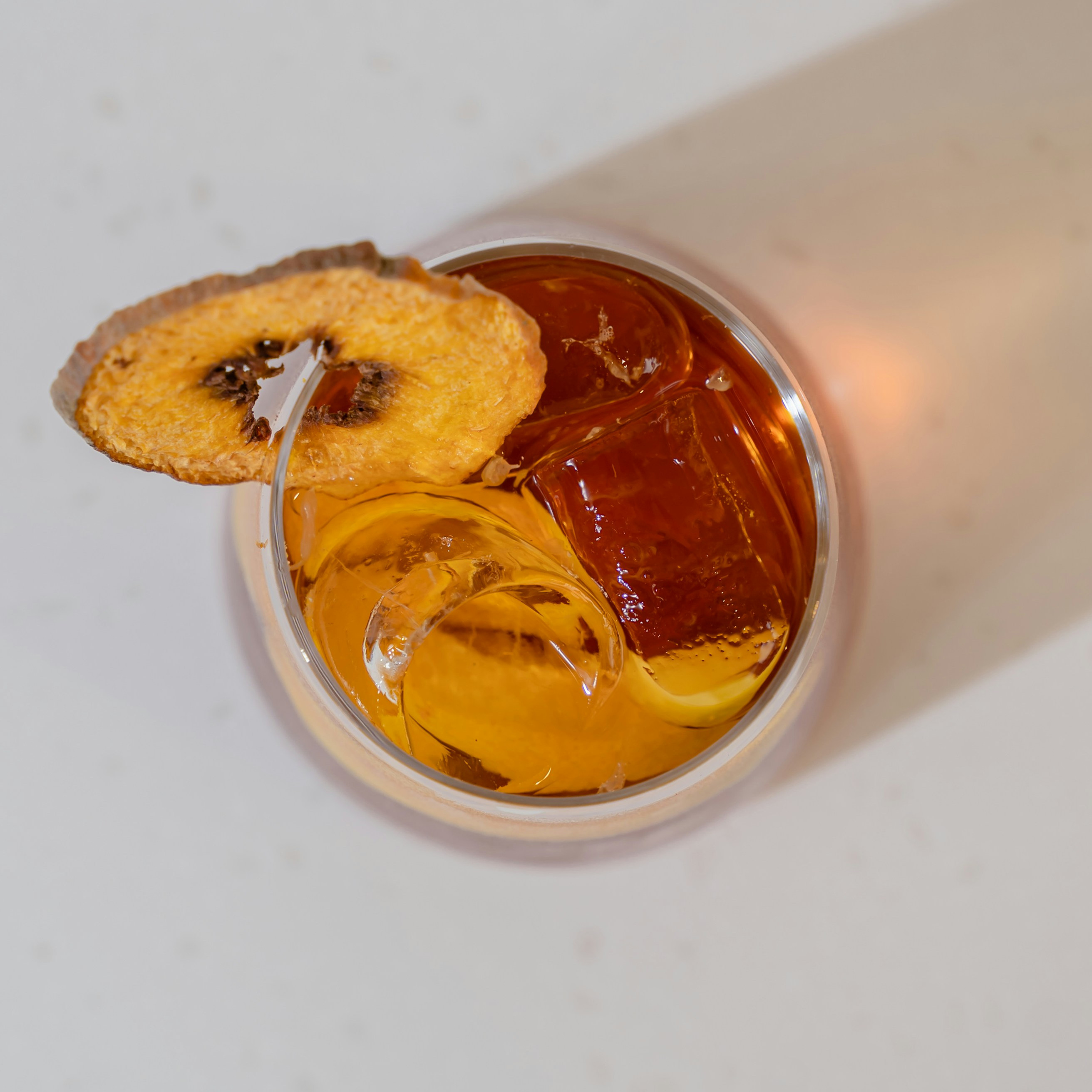 Top view of an amber cocktail with ice and a dried citrus slice on the rim, on a pale table, warm and elegant.