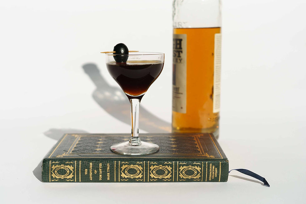 Dark cocktail with an olive garnish sits on a green book, with a liquor bottle blurred behind on a white background.