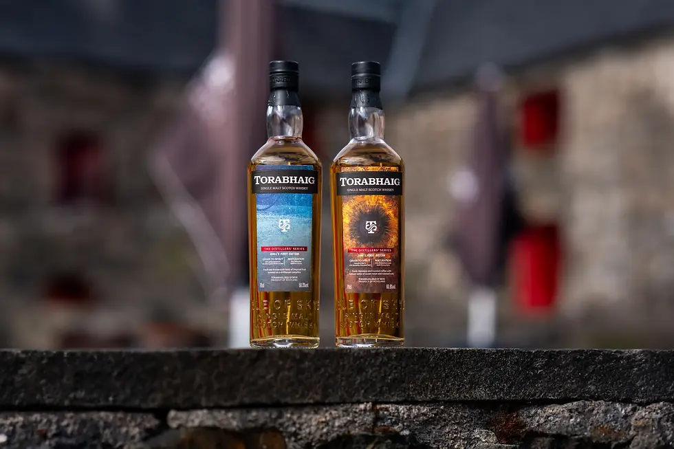 Two Torabhaig single malt Scotch whisky bottles stand on a stone ledge, labels blue and fiery orange, against a blurred distillery background.