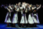 Groupe de danseurs en costumes, bras levés sur scène sombre.