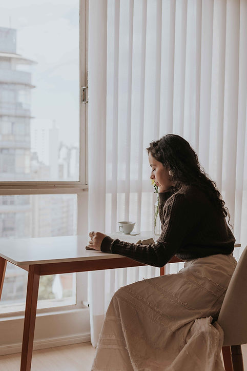 Woman sitting quietly by a window, reflecting — representing the emotional journey of postpartum relationships and motherhood emotional wellbeing