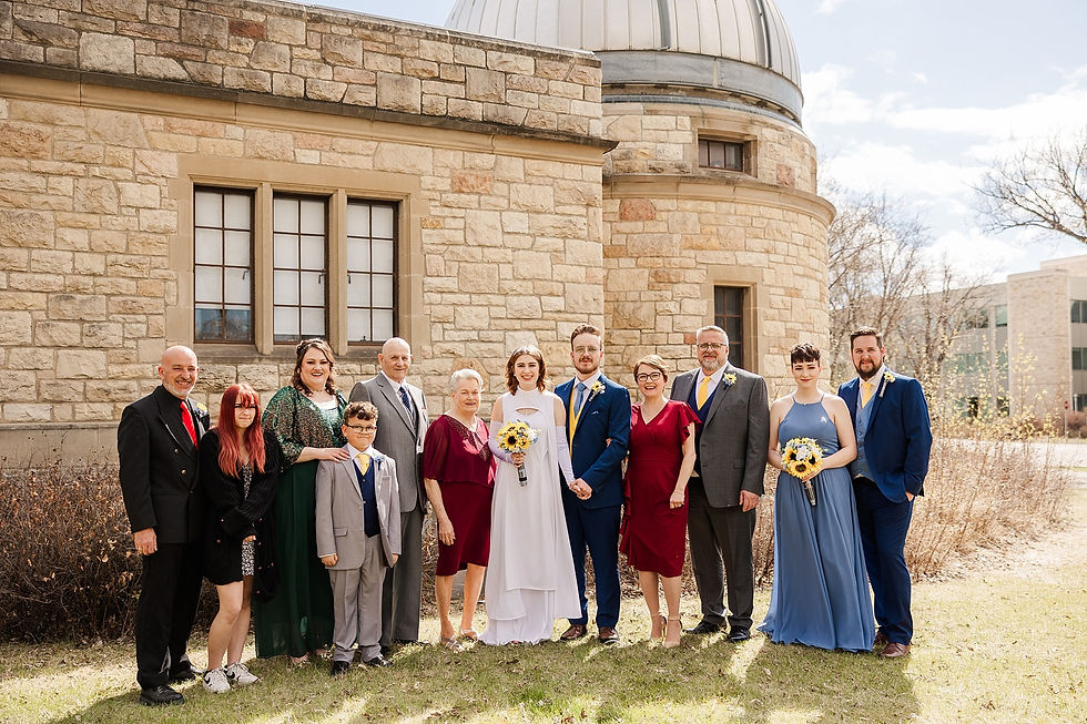 A group of people in formal attire pose smiling in front of a stone building with a dome under a clear sky. They appear happy and celebratory.