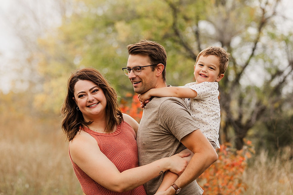 A smiling family of three in a field, with autumn trees in the background. The child is on the man's shoulders, creating a joyful scene.