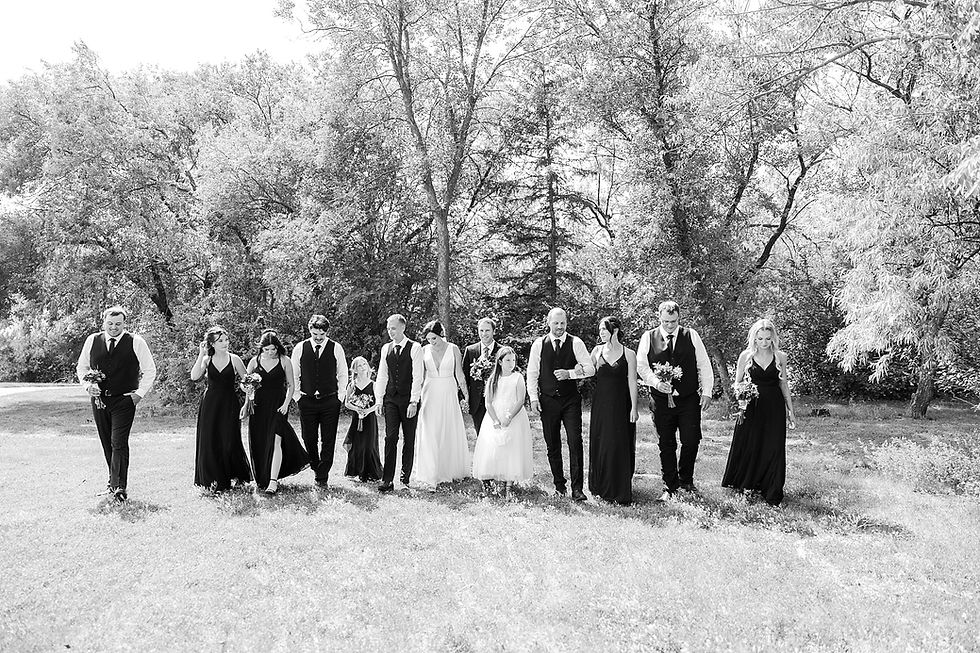 Bridal party walks joyfully through grassy field in black and white photo, surrounded by trees, holding bouquets, dressed in formal attire.