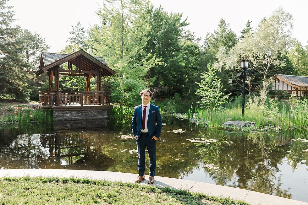 Man in a navy suit stands by a reflective pond near a wooden gazebo and lush trees, calm and formal outdoors.