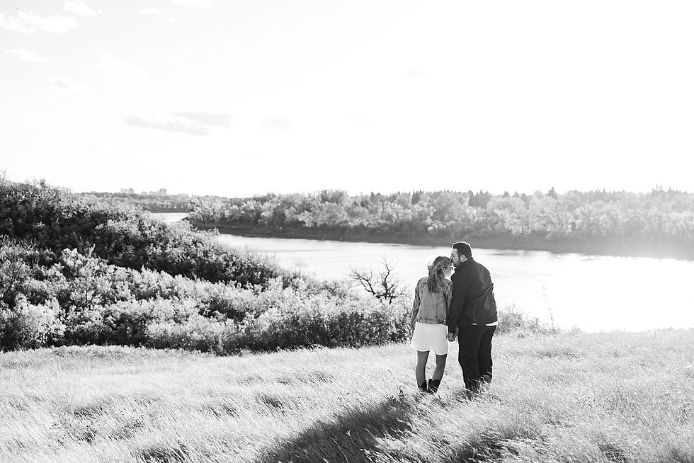 A couple walks hand in hand on a grassy hill overlooking a river, with trees in the background. The scene is in black and white.