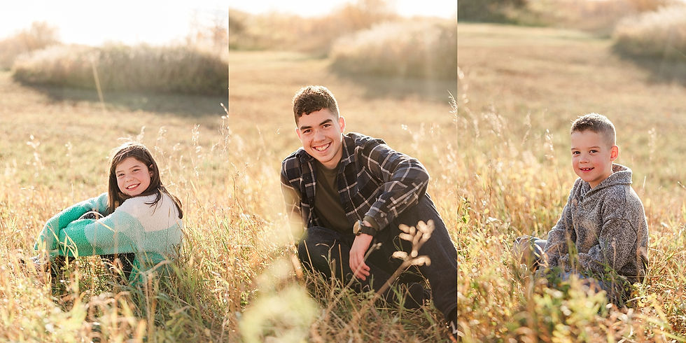 Three children sit and smile in a sunlit field. The girl wears a colorful sweater, the boy a plaid shirt, and the youngest in a gray hoodie.