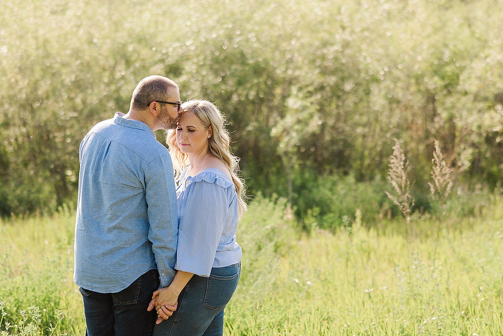A couple in blue shirts lovingly holding hands in a sunlit field. The man kisses the woman's forehead, creating a serene, romantic mood.