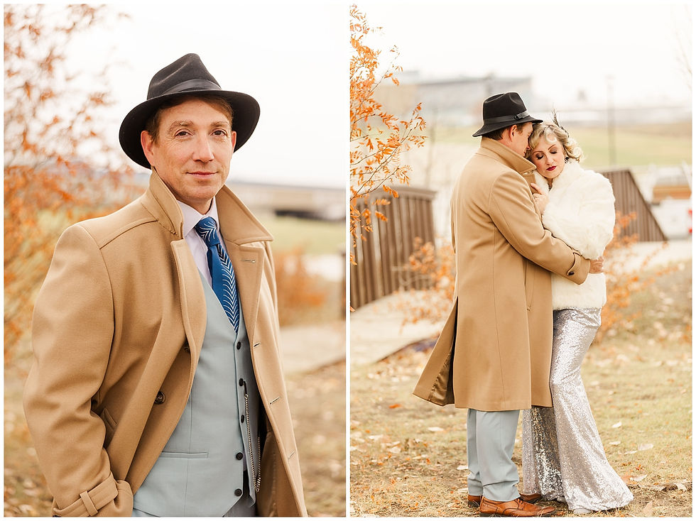 Man in camel coat and hat poses outdoors; hugs woman in white fur and silver dress. Autumn leaves in background, overcast sky.