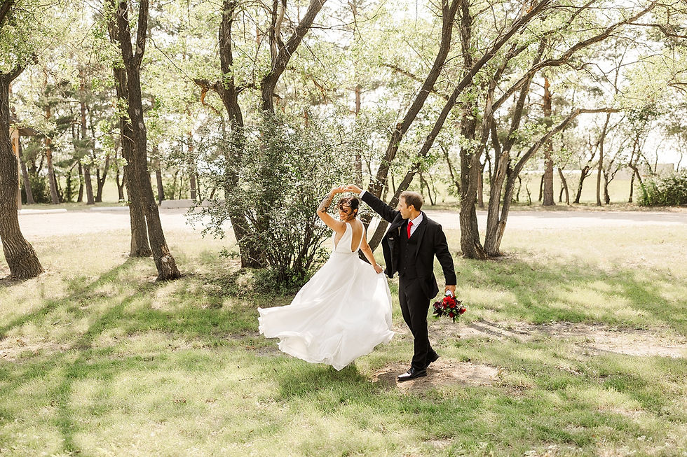 A bride and groom dance joyfully in a sunlit forest, her white dress swirling. He holds red flowers. The mood is romantic and serene.
