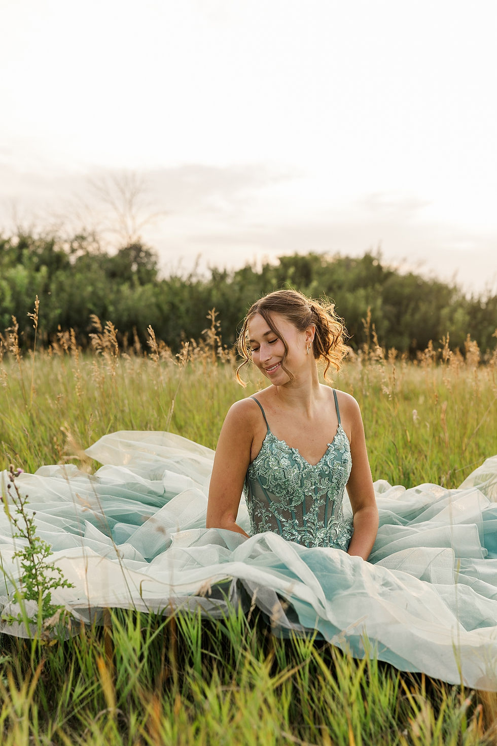 Smiling woman in a teal lace gown sits in a grassy field at sunset, with trees and a bright sky behind her.