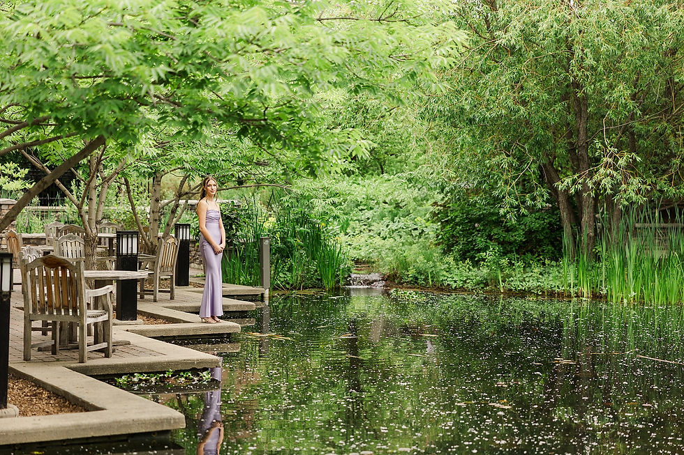 Woman in a lavender dress stands by a pond surrounded by lush greenery and wooden chairs. The peaceful scene reflects in the water.