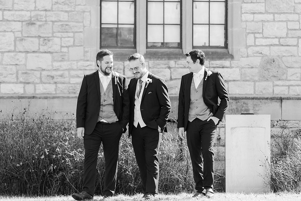 Three men in suits smiling and walking in front of a stone building; wearing boutonnieres, conveying a joyful, celebratory mood.