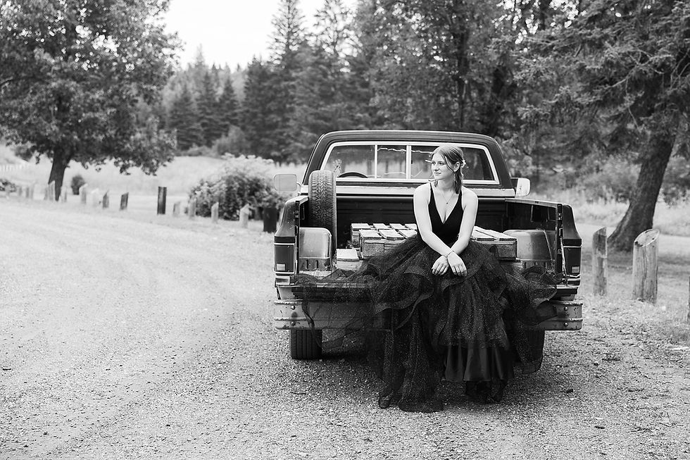 Woman in a black gown sits on a truck bed on a rural gravel road, surrounded by trees. Black and white, contemplative mood. No visible text.