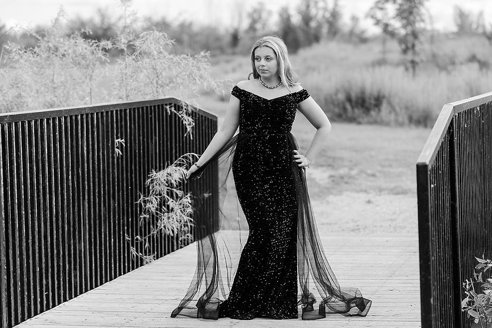Woman in a sparkly black gown stands on a wooden bridge, holding the dress's tulle. Trees and greenery in the blurred background.